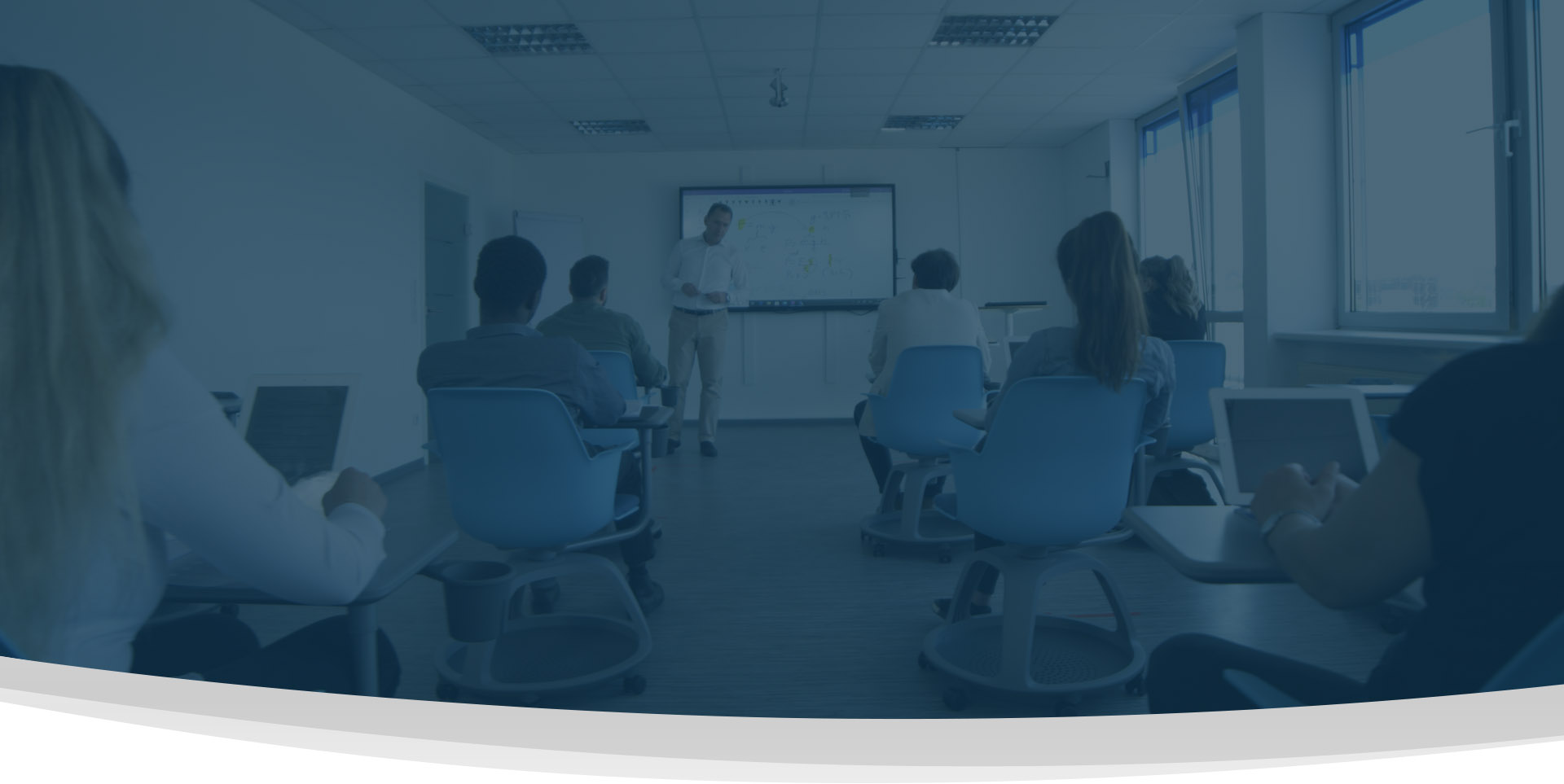 Eine Gruppe von Personen sitzt in einem Klassenzimmer und blickt auf eine Lehrkraft, die in der Nähe eines Bildschirms im vorderen Teil des Raums steht und Inhalte anzeigt. Einige Teilnehmer verwenden Laptops. IFF Akademie - kaufmännische Weiterbildung